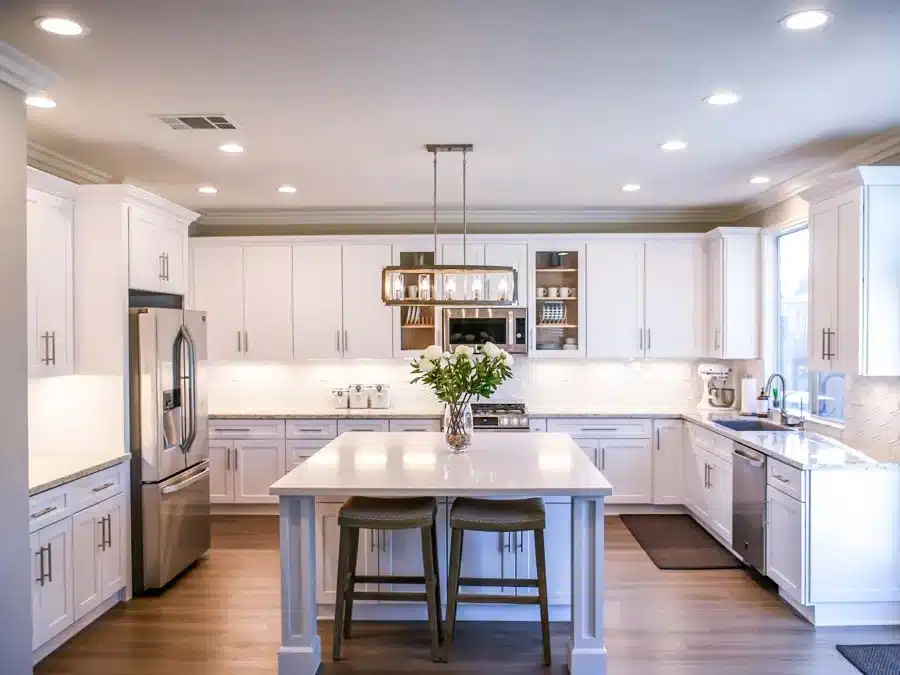 a-kitchen-with-white-cabinets-stainless-steel-appliances-and-a-kitchen-island-with-bar-stools-the-kitchen-has-a-gray-backsplash-and-hardwood-floors-there-is-a-large-window-above-the-sink-that-lets-in-natural-light