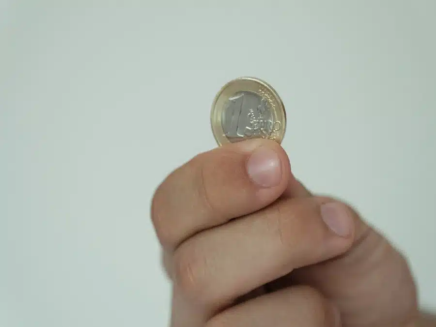close-up of a person's hand holding a gold-colored euro coin between their thumb and forefinger the background is blurred.